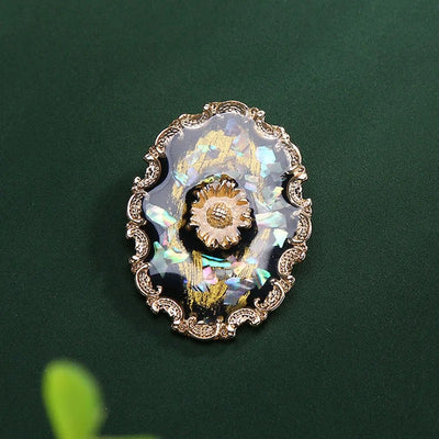 Agatha Resin Cameo Brooch - A round oval-shaped brooch with a black background, with layers of gold enamel paint, abalone shell, and resin, and a pretty stylised sunflower in the centre like a shield.