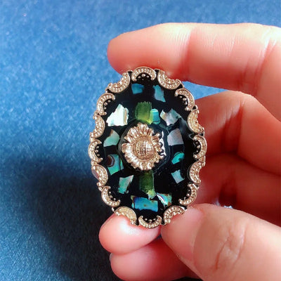 Agatha Resin Cameo Brooch - A round oval-shaped brooch with a black background, with layers of gold enamel paint, abalone shell, and resin, and a pretty stylised sunflower in the centre like a shield.