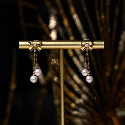 Audrey Bow Tassel Earrings - Tiny, delicate gold earrings shaped like stylised bows, with two tiny pearls suspended from fine gold chains below the lobe.