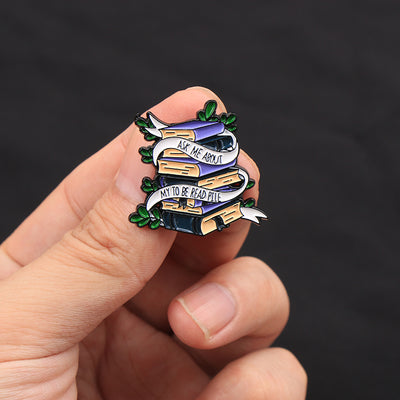 The "Ask Me About My To Be Read Pile" Enamel Pin features a pile of navy blue and purple books wrapped in a ribbon encouraging people to ask about your darkest secret... just how many books are left in your TBR pile. Oh, the shame! The shame! So many books, so little time!
