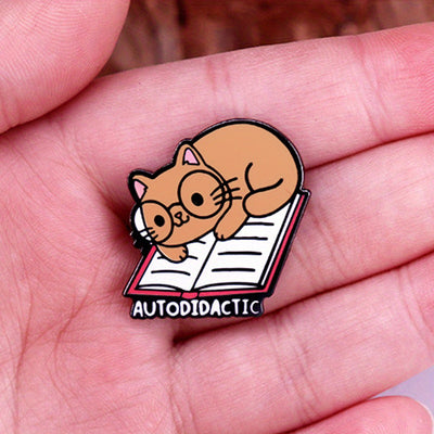 The "Autodidactic" Enamel Pin is a tiny, adorable enamel pin, featuring cute little kitty with big, round glasses reading a book, and the word "Autodidactic" alone the bottom in very small letters.
