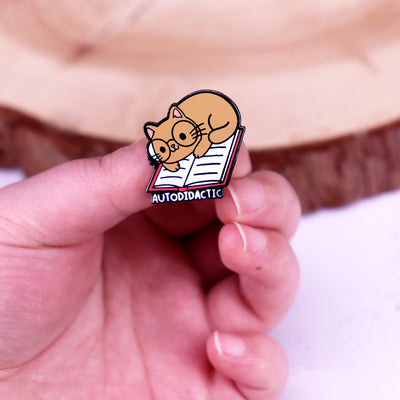 The "Autodidactic" Enamel Pin is a tiny, adorable enamel pin, featuring cute little kitty with big, round glasses reading a book, and the word "Autodidactic" alone the bottom in very small letters.