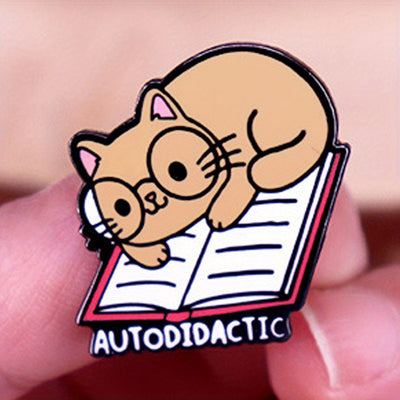 The "Autodidactic" Enamel Pin is a tiny, adorable enamel pin, featuring cute little kitty with big, round glasses reading a book, and the word "Autodidactic" alone the bottom in very small letters.