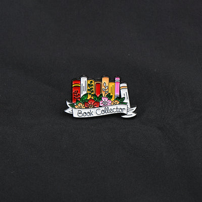 The "Book Collector" Enamel Pin is an adorable, teeny-tiny little enamel pin, featuring a row of stylised books in shades of pink, orange, and red, adorned with flowers and ribbons. So cute! Grab yours today!