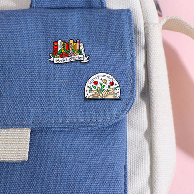 The "Book Collector" Enamel Pin is an adorable, teeny-tiny little enamel pin, featuring a row of stylised books in shades of pink, orange, and red, adorned with flowers and ribbons. So cute! Grab yours today!