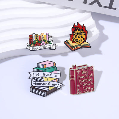 The "Book Collector" Enamel Pin is an adorable, teeny-tiny little enamel pin, featuring a row of stylised books in shades of pink, orange, and red, adorned with flowers and ribbons. So cute! Grab yours today!