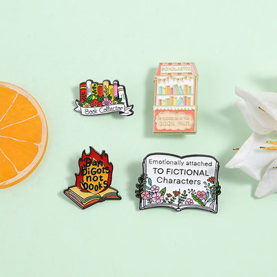 The "Book Collector" Enamel Pin is an adorable, teeny-tiny little enamel pin, featuring a row of stylised books in shades of pink, orange, and red, adorned with flowers and ribbons. So cute! Grab yours today!