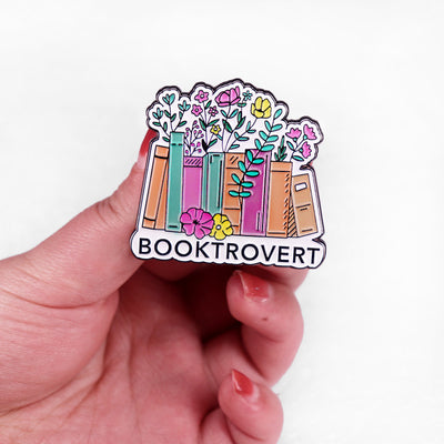 The "Booktrovert" Enamel Pin is a large pin featuring a row of colourful books, adorned with stylised flowers and leaves, with the slogan along the bottom like a bookshelf. Simple, colourful, and lovely! Grab yours today!