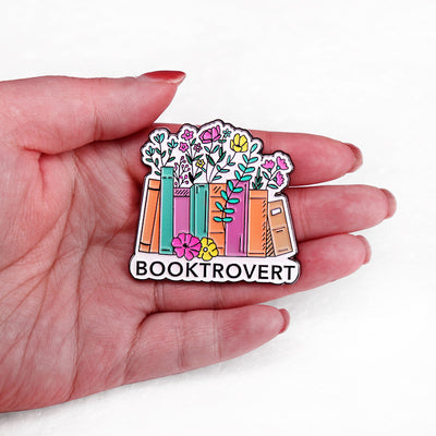 The "Booktrovert" Enamel Pin is a large pin featuring a row of colourful books, adorned with stylised flowers and leaves, with the slogan along the bottom like a bookshelf. Simple, colourful, and lovely! Grab yours today!