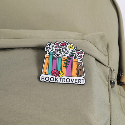 The "Booktrovert" Enamel Pin is a large pin featuring a row of colourful books, adorned with stylised flowers and leaves, with the slogan along the bottom like a bookshelf. Simple, colourful, and lovely! Grab yours today!