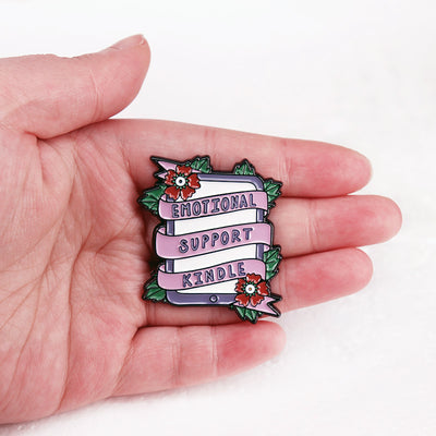 The "Emotional Support Kindle" Enamel Pin features a stylised Kindle eBook reader, adorned with a pink ribbon, green leaves, and red flowers in the corners.