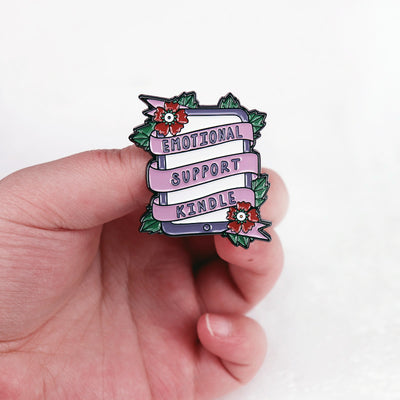 The "Emotional Support Kindle" Enamel Pin features a stylised Kindle eBook reader, adorned with a pink ribbon, green leaves, and red flowers in the corners.