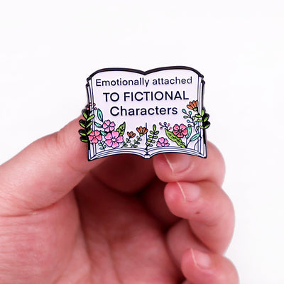 The "Emotionally Attached To Fictional Characters" Enamel Pin features a stylised book adorned with delicate leaves and flowers, and text proclaiming your love of certain fictional characters. Oh my! No judgement here!