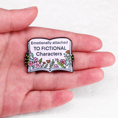 The "Emotionally Attached To Fictional Characters" Enamel Pin features a stylised book adorned with delicate leaves and flowers, and text proclaiming your love of certain fictional characters. Oh my! No judgement here!