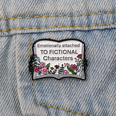 The "Emotionally Attached To Fictional Characters" Enamel Pin features a stylised book adorned with delicate leaves and flowers, and text proclaiming your love of certain fictional characters. Oh my! No judgement here!