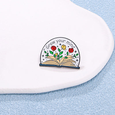 The "Grow Your Mind" Enamel Pin an average-sized brooch featuring a stylised book with flowers and vines sprouting from the pages, and text encouraging you to grow your mind. So true!