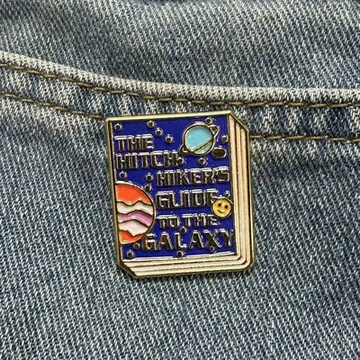 The "Hitchhiker's Guider" Enamel Pin features a stylised replica of the cult classic book, The Hitchhiker's Guide To The Galaxy, in vibrant coloured enamel paint, edged with metallic gold.