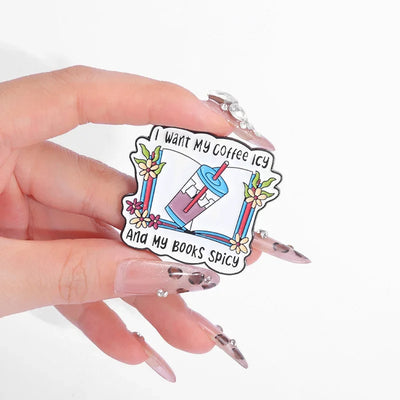 The "I Want My Coffee Icy And My Books Spicy" Enamel Pin features a stylised cup of iced coffee atop a book, adorned with colourful flowers and leaves.