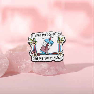 The "I Want My Coffee Icy And My Books Spicy" Enamel Pin features a stylised cup of iced coffee atop a book, adorned with colourful flowers and leaves.