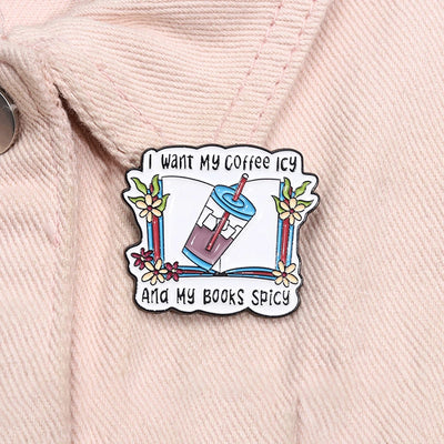 The "I Want My Coffee Icy And My Books Spicy" Enamel Pin features a stylised cup of iced coffee atop a book, adorned with colourful flowers and leaves.