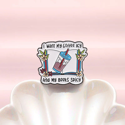 The "I Want My Coffee Icy And My Books Spicy" Enamel Pin features a stylised cup of iced coffee atop a book, adorned with colourful flowers and leaves.