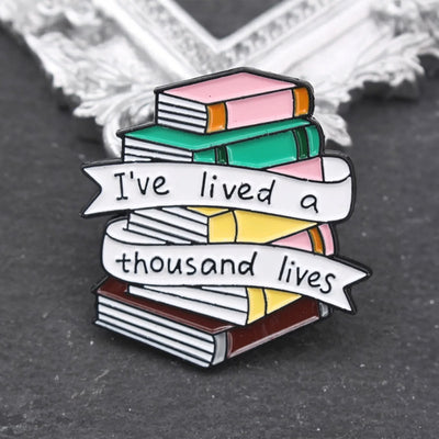The "I've Lived A Thousand Lives" Enamel Pin features a stack of colourful stylised cartoon books, with a ribbon on the front announcing just how many fantastical lives you've lived. At least a thousand! Maybe more!