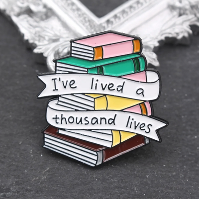 The "I've Lived A Thousand Lives" Enamel Pin features a stack of colourful stylised cartoon books, with a ribbon on the front announcing just how many fantastical lives you've lived. At least a thousand! Maybe more!
