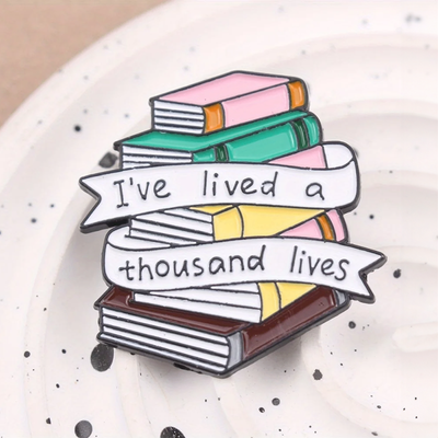 The "I've Lived A Thousand Lives" Enamel Pin features a stack of colourful stylised cartoon books, with a ribbon on the front announcing just how many fantastical lives you've lived. At least a thousand! Maybe more!