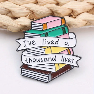 The "I've Lived A Thousand Lives" Enamel Pin features a stack of colourful stylised cartoon books, with a ribbon on the front announcing just how many fantastical lives you've lived. At least a thousand! Maybe more!