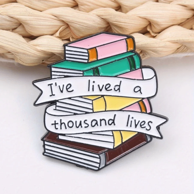 The "I've Lived A Thousand Lives" Enamel Pin features a stack of colourful stylised cartoon books, with a ribbon on the front announcing just how many fantastical lives you've lived. At least a thousand! Maybe more!