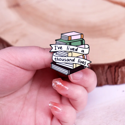 The "I've Lived A Thousand Lives" Enamel Pin features a stack of colourful stylised cartoon books, with a ribbon on the front announcing just how many fantastical lives you've lived. At least a thousand! Maybe more!