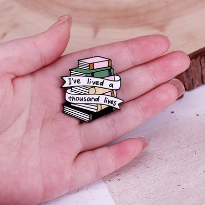 The "I've Lived A Thousand Lives" Enamel Pin features a stack of colourful stylised cartoon books, with a ribbon on the front announcing just how many fantastical lives you've lived. At least a thousand! Maybe more!