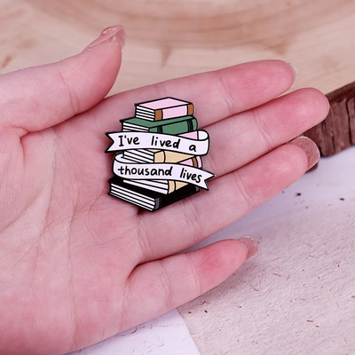 The "I've Lived A Thousand Lives" Enamel Pin features a stack of colourful stylised cartoon books, with a ribbon on the front announcing just how many fantastical lives you've lived. At least a thousand! Maybe more!