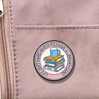 The "Life Isn't Hard When You Have A Library Card" Enamel Pin features a stack of books in the centre (plus a cup of sweet, sweet coffee!), within a ring of text proclaiming your love for libraries.