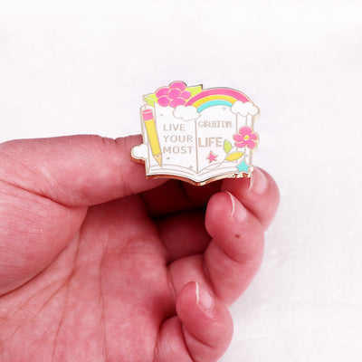 The "Live Your Most Creative Life" Enamel Pin is an average-sized brooch featuring a stylised book, decorated with flowers, rainbow, and art supplies. Perfect for the creative soul!