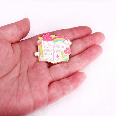 The "Live Your Most Creative Life" Enamel Pin is an average-sized brooch featuring a stylised book, decorated with flowers, rainbow, and art supplies. Perfect for the creative soul!