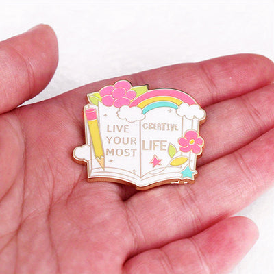 The "Live Your Most Creative Life" Enamel Pin is an average-sized brooch featuring a stylised book, decorated with flowers, rainbow, and art supplies. Perfect for the creative soul!