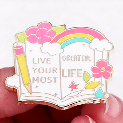 The "Live Your Most Creative Life" Enamel Pin is an average-sized brooch featuring a stylised book, decorated with flowers, rainbow, and art supplies. Perfect for the creative soul!