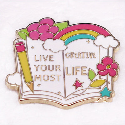 The "Live Your Most Creative Life" Enamel Pin is an average-sized brooch featuring a stylised book, decorated with flowers, rainbow, and art supplies. Perfect for the creative soul!