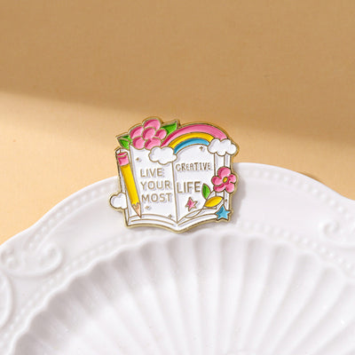 The "Live Your Most Creative Life" Enamel Pin is an average-sized brooch featuring a stylised book, decorated with flowers, rainbow, and art supplies. Perfect for the creative soul!