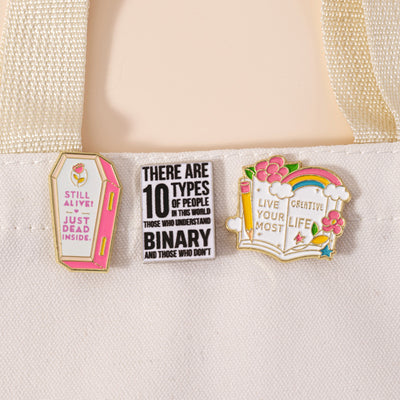 The "Live Your Most Creative Life" Enamel Pin is an average-sized brooch featuring a stylised book, decorated with flowers, rainbow, and art supplies. Perfect for the creative soul!