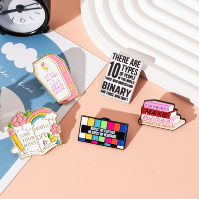 The "Live Your Most Creative Life" Enamel Pin is an average-sized brooch featuring a stylised book, decorated with flowers, rainbow, and art supplies. Perfect for the creative soul!