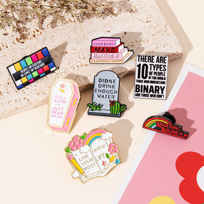 The "Live Your Most Creative Life" Enamel Pin is an average-sized brooch featuring a stylised book, decorated with flowers, rainbow, and art supplies. Perfect for the creative soul!