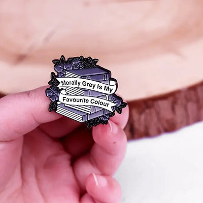 The "Morally Grey Is My Favourite Colour" Enamel Pin cool, stylised pin featuring a stack of stylised books with different shades of grey covers and a ribbon woven around them featuring the slogan.