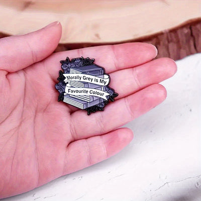 The "Morally Grey Is My Favourite Colour" Enamel Pin cool, stylised pin featuring a stack of stylised books with different shades of grey covers and a ribbon woven around them featuring the slogan.