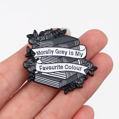 The "Morally Grey Is My Favourite Colour" Enamel Pin cool, stylised pin featuring a stack of stylised books with different shades of grey covers and a ribbon woven around them featuring the slogan.