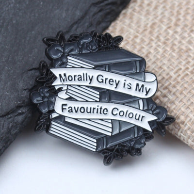 The "Morally Grey Is My Favourite Colour" Enamel Pin cool, stylised pin featuring a stack of stylised books with different shades of grey covers and a ribbon woven around them featuring the slogan.