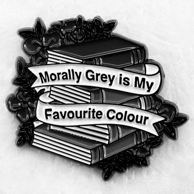 The "Morally Grey Is My Favourite Colour" Enamel Pin cool, stylised pin featuring a stack of stylised books with different shades of grey covers and a ribbon woven around them featuring the slogan.