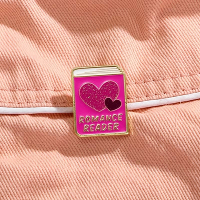 The "Romance Reader" Enamel Pin is a simple but elegant design, featuring a pink book outlined in gold, adorned with hearts. Perfect for expressing your love of the romance genre to all. Grab yours today!