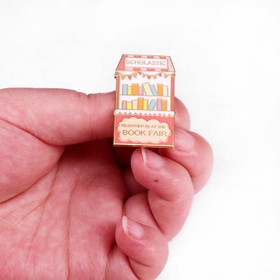 The "Scholastic Book Fair" Enamel Pin features a stylised bookshelf in lovely pastel shades of pink, coral, and gold, commemorating everyone's favourite childhood memory: The Scholastic Book Fair! Oh, the joy! So many books!
